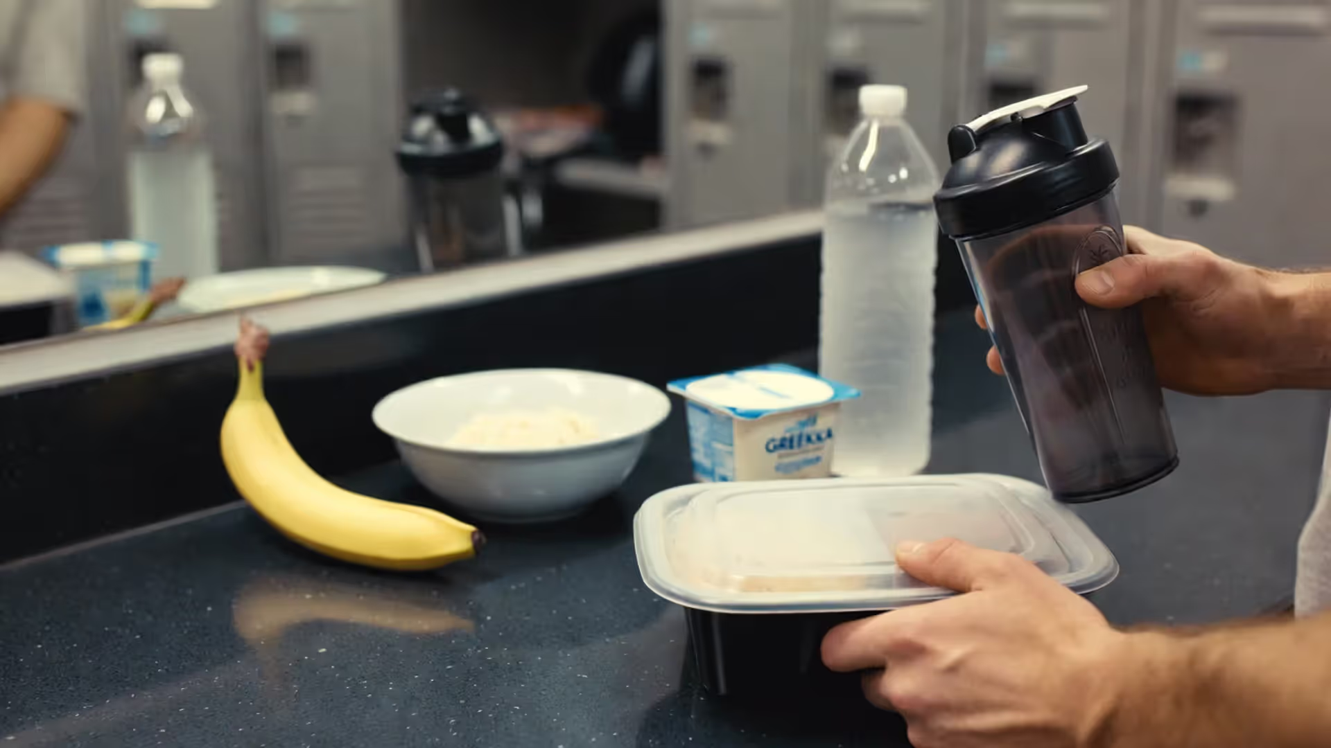 Person holding a protein shake and a meal container with common post-workout foods on a counter.