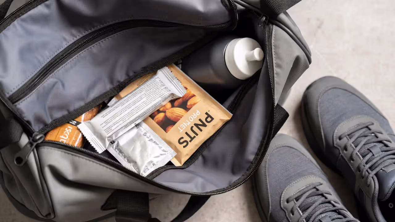 Portable fitness snacks in a gym bag including protein bar, nuts, nut butter packet, and water bottle